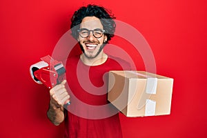 Handsome hispanic man holding packing tape and cardboard box smiling and laughing hard out loud because funny crazy joke