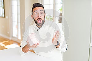 Handsome hispanic man holding piggy bank very happy and excited, winner expression celebrating victory screaming with big smile