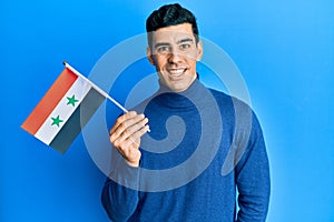 Handsome hispanic man holding syria flag looking positive and happy standing and smiling with a confident smile showing teeth