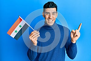 Handsome hispanic man holding syria flag smiling happy pointing with hand and finger to the side