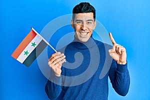 Handsome hispanic man holding syria flag smiling with an idea or question pointing finger with happy face, number one
