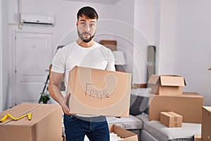 Handsome hispanic man moving to a new home holding fragile box clueless and confused expression