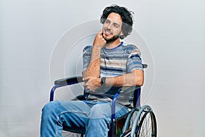 Handsome hispanic man sitting on wheelchair with hand on chin thinking about question, pensive expression