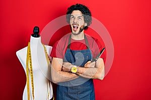 Handsome hispanic man standing by manikin holding scissors angry and mad screaming frustrated and furious, shouting with anger