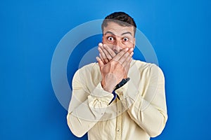 Handsome hispanic man standing over blue background shocked covering mouth with hands for mistake