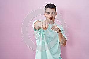 Handsome hispanic man standing over pink background punching fist to fight, aggressive and angry attack, threat and violence