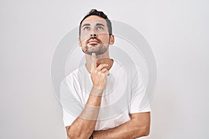Handsome hispanic man standing over white background thinking concentrated about doubt with finger on chin and looking up