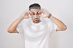 Handsome hispanic man standing over white background trying to open eyes with fingers, sleepy and tired for morning fatigue