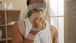 Handsome hispanic man using a jade roller in a tranquil spa room with soft lighting and minimalist decor elements surrounding the photo