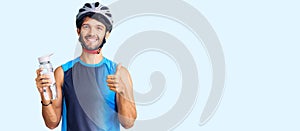 Handsome hispanic man wearing bike helmet and holding water bottle smiling happy and positive, thumb up doing excellent and