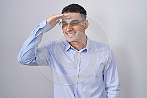 Handsome hispanic man wearing business clothes and glasses very happy and smiling looking far away with hand over head