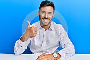 Handsome hispanic man wearing business clothes sitting on the table doing happy thumbs up gesture with hand