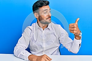 Handsome hispanic man wearing business clothes sitting on the table looking proud, smiling doing thumbs up gesture to the side