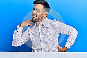 Handsome hispanic man wearing business clothes sitting on the table suffering of neck ache injury, touching neck with hand, photo