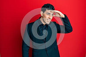 Handsome hispanic man wearing business clothes very happy and smiling looking far away with hand over head