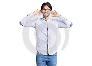 Handsome hispanic man wearing business shirt and glasses trying to hear both hands on ear gesture, curious for gossip