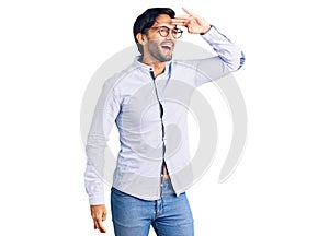 Handsome hispanic man wearing business shirt and glasses very happy and smiling looking far away with hand over head