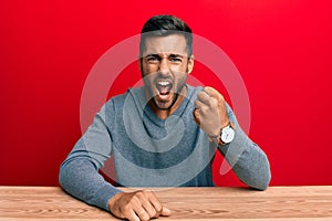 Handsome hispanic man wearing casual style sitting on the table angry and mad raising fist frustrated and furious while shouting