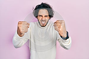 Handsome hispanic man wearing casual white sweater angry and mad raising fists frustrated and furious while shouting with anger
