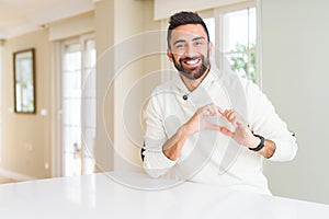 Handsome hispanic man wearing casual white sweater at home smiling in love showing heart symbol and shape with hands