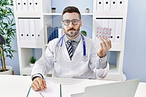 Handsome hispanic man wearing doctor uniform holding prescription pills thinking attitude and sober expression looking self