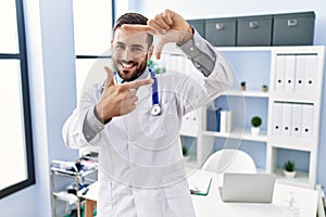 Handsome hispanic man wearing doctor uniform and stethoscope at medical clinic smiling making frame with hands and fingers with
