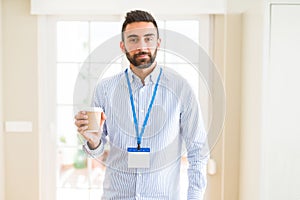 Handsome hispanic man wearing id card and drinking a cup of coffee with a confident expression on smart face thinking serious