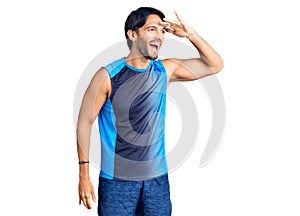 Handsome hispanic man wearing sportswear very happy and smiling looking far away with hand over head