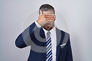 Handsome hispanic man wearing suit and tie covering eyes with hand, looking serious and sad