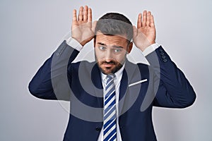 Handsome hispanic man wearing suit and tie doing bunny ears gesture with hands palms looking cynical and skeptical