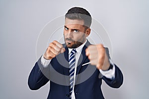Handsome hispanic man wearing suit and tie punching fist to fight, aggressive and angry attack, threat and violence
