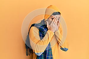 Handsome hispanic man wearing wool hat and winter scarf with sad expression covering face with hands while crying