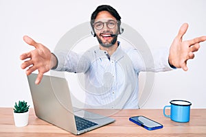 Handsome hispanic man working at the office wearing operator headset looking at the camera smiling with open arms for hug