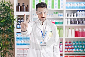 Handsome hispanic man working at pharmacy drugstore showing and pointing up with fingers number two while smiling confident and