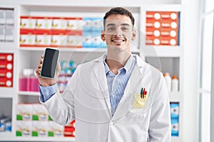Handsome hispanic man working at pharmacy drugstore showing smartphone screen looking positive and happy standing and smiling with