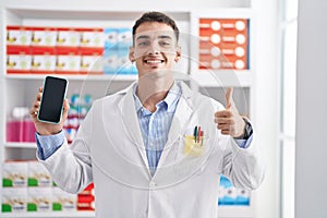 Handsome hispanic man working at pharmacy drugstore showing smartphone screen smiling happy and positive, thumb up doing excellent