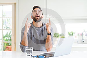 Handsome hispanic man working using computer laptop amazed and surprised looking up and pointing with fingers and raised arms