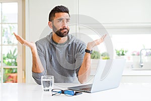 Handsome hispanic man working using computer laptop clueless and confused expression with arms and hands raised
