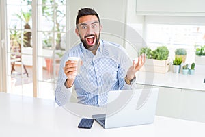 Handsome hispanic man working using computer laptop and drinking a cup of coffee very happy and excited, winner expression