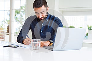 Handsome hispanic man working using computer and writing on a paper with a confident expression on smart face thinking serious