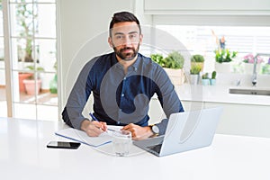 Handsome hispanic man working using computer and writing on a paper with a confident expression on smart face thinking serious