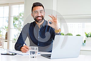 Handsome hispanic man working using computer and writing on a paper doing ok sign with fingers, excellent symbol