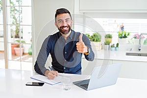 Handsome hispanic man working using computer and writing on a paper happy with big smile doing ok sign, thumb up with fingers,