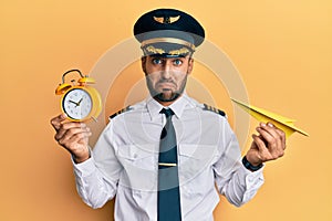 Handsome hispanic pilot man holding paper plane and alarm clock depressed and worry for distress, crying angry and afraid