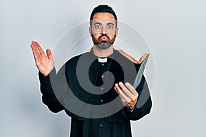 Handsome hispanic priest man with beard holding bible and christian cross puffing cheeks with funny face