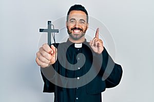 Handsome hispanic priest man with beard holding catholic cross surprised with an idea or question pointing finger with happy face,