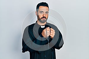 Handsome hispanic priest man with beard praying holding catholic rosary relaxed with serious expression on face