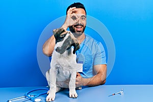 Handsome hispanic veterinary man with beard checking dog health doing ok gesture with hand smiling, eye looking through fingers