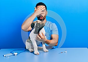 Handsome hispanic veterinary man with beard checking dog health peeking in shock covering face and eyes with hand, looking through