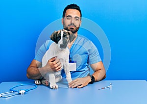 Handsome hispanic veterinary man with beard checking dog health relaxed with serious expression on face